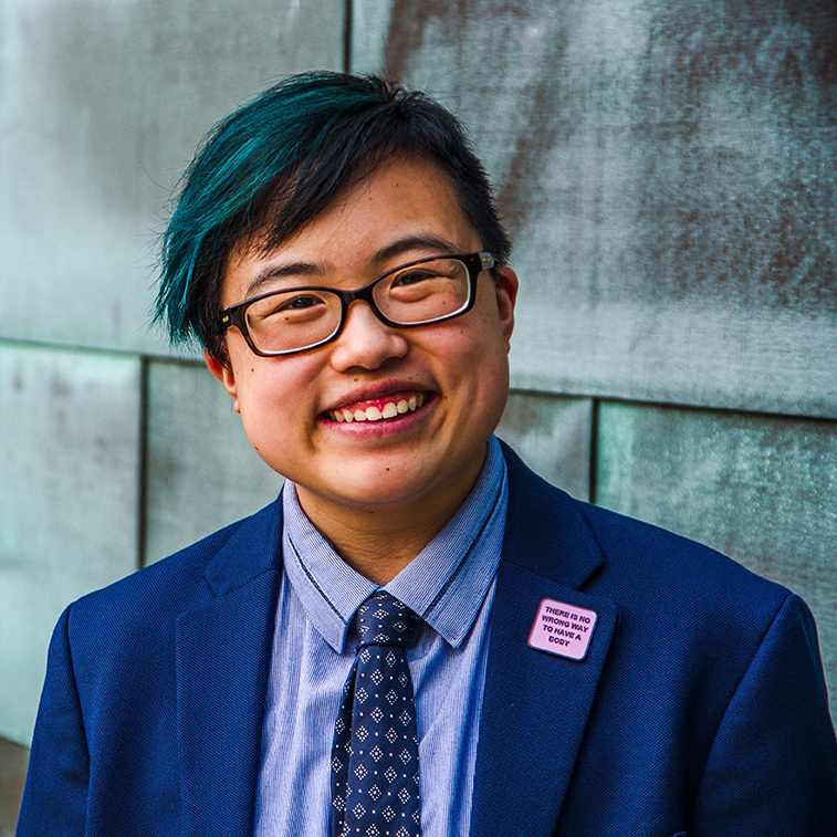 [Photo: Lydia smiles and tilts their head slightly to the side, looking confidently at the camera. They are a young-ish East Asian person with a streak of teal in their short black hair, wearing glasses, a cobalt blue jacket and navy tie, with a blue copper wall behind them. Photo by Sarah Tundermann.]